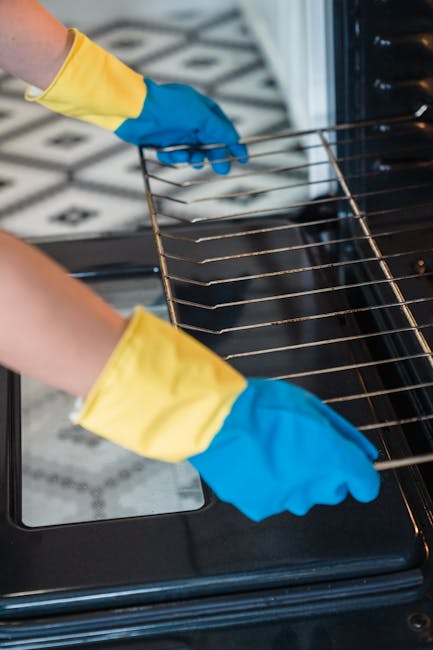 Close-up of a person wearing yellow and blue cleaning gloves, inserting a wire rack into a modern black oven with a glass door. The oven interior appears clean and shiny, while the surrounding counter